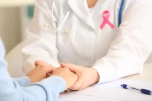 Lekarz wspiera pacjentkę trzymając ją za dłonie, na fartuchu widać różową wstążkę symbolizującą walkę z rakiem piersi